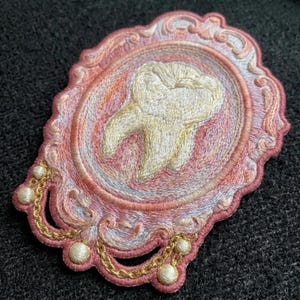 May include: An embroidered patch featuring a central cream-colored tooth motif encased within an ornate, oval-shaped pink and peach frame. The design includes decorative gold-colored thread chains and small white spherical accents along the border.