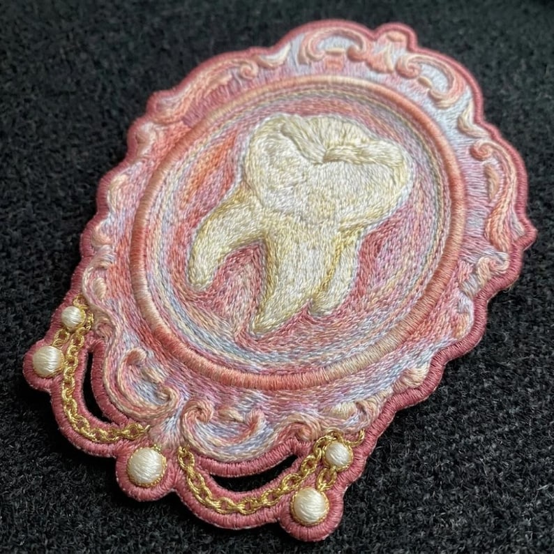 May include: An embroidered patch featuring a central cream-colored tooth motif encased within an ornate, oval-shaped pink and peach frame. The design includes decorative gold-colored thread chains and small white spherical accents along the border.