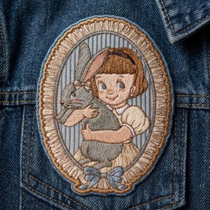 May include: An oval embroidered patch featuring a child with brown hair holding a gray rabbit. The design is framed by a tan ruffled border and a blue striped background, with a blue bow at the bottom, all stitched onto a blue denim fabric.