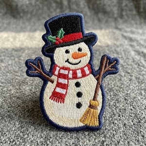 May include: An embroidered patch featuring a snowman with a black top hat, a red and white striped scarf, and a carrot nose. The snowman has three black buttons on its body, brown stick arms, and holds a yellow broom. The patch has a dark blue border.