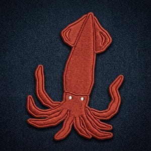 May include: An embroidered patch in the shape of a squid, featuring a deep rust-orange thread color. The design includes a long, pointed mantle, two small white circular eyes, and multiple curved tentacles extending outward from the central body.