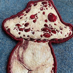 May include: An embroidered patch shaped like a mushroom with a cream-colored cap featuring dark red spots. The mushroom stem is cream-colored with subtle shading, and the entire design is outlined with a thick, dark red stitched border.