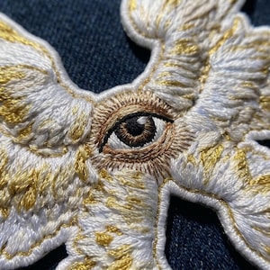 May include: An embroidered patch featuring a central human eye with a brown iris and black pupil, surrounded by detailed white and metallic gold wings. The stitching creates a textured, layered effect with white thread and gold accents.