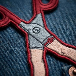 May include: An embroidered patch featuring a pair of scissors with handles shaped like human legs wearing light blue shorts. The scissors are outlined in thick red thread and are stitched onto a dark blue denim fabric background.