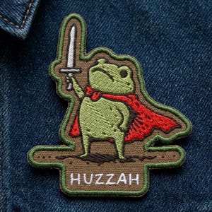 May include: An embroidered patch featuring a light green frog wearing a red cape and holding a silver sword. The frog stands on a brown base with the word HUZZAH stitched in white letters. The patch has a dark green border and is textured.