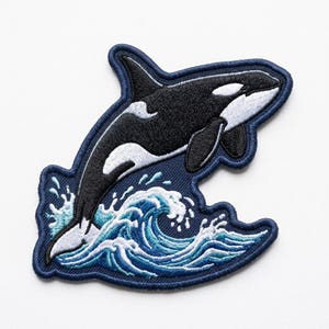 May include: An embroidered patch featuring a black and white orca whale leaping out of a stylized blue and white ocean wave. The design is outlined with a dark blue border and showcases detailed stitching of the whale and splashing water.