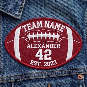 May include: An oval-shaped maroon embroidered patch featuring a white football graphic, the text TEAM NAME, ALEXANDER, 42, and EST. 2023. The patch is stitched onto a blue denim fabric background with a visible copper-colored metal button.