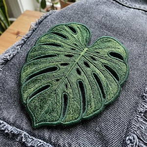 May include: A green embroidered monstera leaf patch attached to a dark gray denim fabric. The patch features detailed stitching in various shades of green, capturing the characteristic fenestrations and heart-shaped outline of the tropical plant leaf.