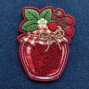 May include: An embroidered patch featuring a jar of red strawberry jam. The jar lid is covered with a red and white checkered cloth, tied with a tan string bow. Green leaves, a white flower, and three red strawberries sit atop the lid.