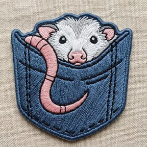 May include: An embroidered patch featuring a white and gray opossum with a pink nose and ears peeking out from a blue denim pocket. A long, curved pink tail extends from the pocket, wrapping around the front of the design with detailed stitching.