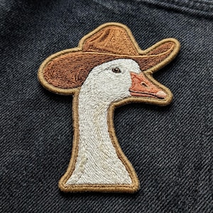 May include: An embroidered patch featuring a white goose wearing a brown cowboy hat. The goose has a light orange beak and is depicted in profile facing right. The patch is stitched with detailed thread work and has a brown border outline.