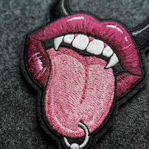 May include: An embroidered patch featuring a pair of dark pink lips with a light pink tongue sticking out. The design includes sharp white fangs, a silver-colored ring piercing the tongue, and a black border outline on a dark gray fabric background.