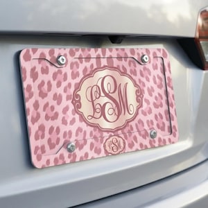 May include: Pink license plate with a leopard print pattern. The plate features a central monogram in a decorative frame, with the initials "LSM". A smaller version of the monogram is at the bottom. The plate is attached to a vehicle with four screws.