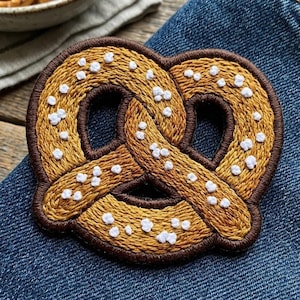May include: A pretzel-shaped embroidered patch featuring golden-brown thread with a dark brown border and small, raised white stitched accents representing salt crystals. The patch has a classic knotted design with three distinct open loops.