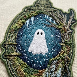 May include: An embroidered patch featuring a white ghost centered within a dark blue, star-filled oval frame. The border is shaped like an ornate, dark green frame decorated with textured mossy elements and bare, brown tree branches.