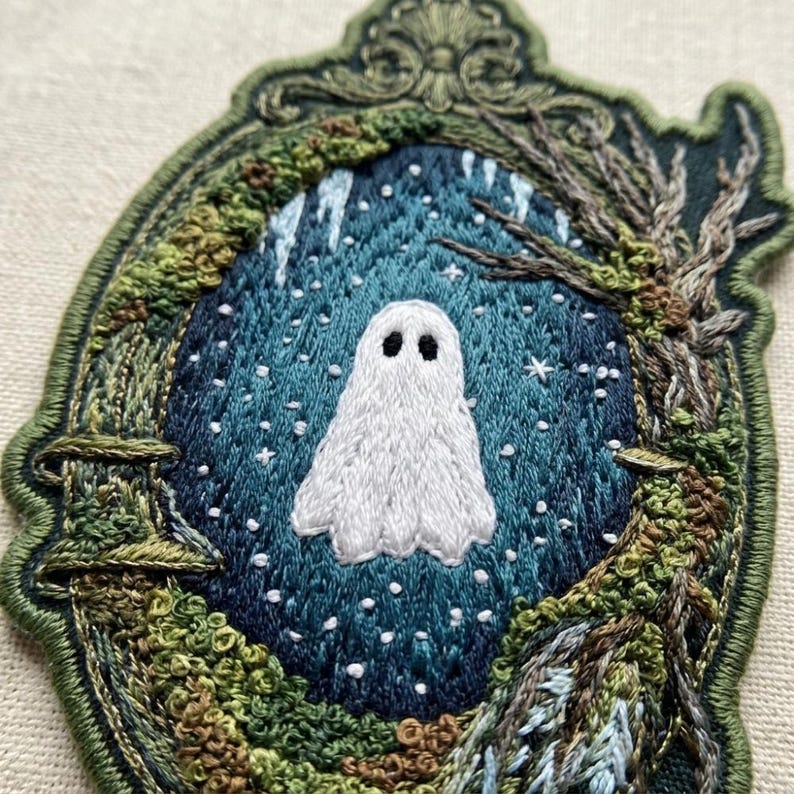 May include: An embroidered patch featuring a white ghost centered within a dark blue, star-filled oval frame. The border is shaped like an ornate, dark green frame decorated with textured mossy elements and bare, brown tree branches.