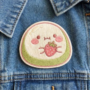 May include: A circular embroidered patch featuring a cream-colored character with pink cheeks and a small mouth, holding a red strawberry with green leaves. The patch has a light green border and is attached to a blue denim fabric background.