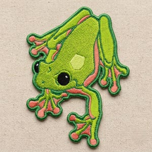 May include: An embroidered patch featuring a bright green frog with large black eyes and coral-pink accents on its toes and sides. The frog is depicted in a dynamic, sprawling pose with its limbs extended, showcasing detailed stitching throughout.