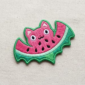May include: An embroidered patch shaped like a bat with a watermelon-inspired design. The central body is pink with black seed-like markings, while the wings are pink with a green border, featuring two white eyes and pointed ears.