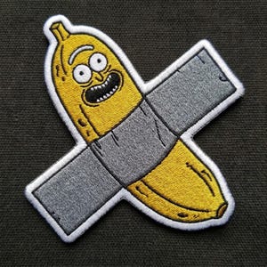 May include: An embroidered patch featuring a yellow banana with a cartoon face, including large white eyes with black pupils and an open mouth with teeth, partially covered by a wide, horizontal strip of grey duct tape with black outlines.