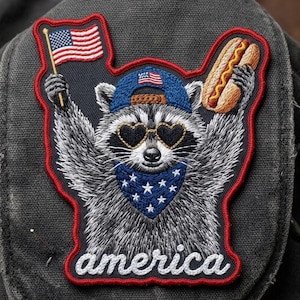 May include: An embroidered patch featuring a raccoon wearing a blue baseball cap with a small American flag, heart-shaped sunglasses, and a blue bandana with white stars. The raccoon holds an American flag and a hot dog. The word america is below.