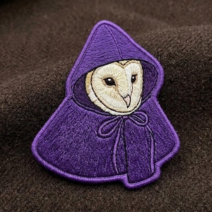 May include: An embroidered patch featuring a barn owl with a cream and tan face, wearing a deep purple hooded cloak. The cloak has a pointed hood and a tied front closure with visible stitching detail throughout the purple fabric and owl face.