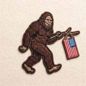 May include: An embroidered patch featuring a brown, furry Bigfoot creature walking while holding a wooden stick. Hanging from the stick is a small, rectangular American flag with red and white stripes and a blue field with white stars.