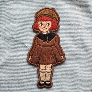 May include: An embroidered patch featuring a character with bright red hair, a brown beret, and a brown dress with a black collar and bow. The character has freckles and wears light beige knee-high socks with dark shoes, rendered in detailed thread.