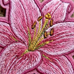 May include: An embroidered patch featuring a pink lily flower with white highlights and yellow stamens. The petals are stitched with detailed thread work in varying shades of pink, creating a textured appearance with a dark pink border.