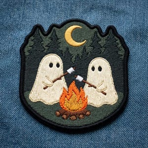 May include: An embroidered patch featuring two off-white ghosts sitting by a campfire with orange and yellow flames. The ghosts hold brown sticks with white marshmallows. A yellow crescent moon shines above a dark green forest background.