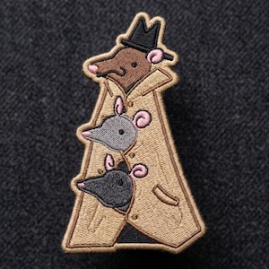 May include: An embroidered patch featuring three small rodents stacked vertically inside a tan trench coat. The top rodent wears a black fedora hat. Each rodent has a pointed snout, small pink ears, and black eyes. The design is stitched with tan, brown, gray, black, and pink thread.