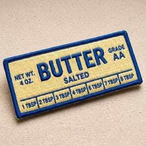 May include: A rectangular embroidered patch featuring a yellow background with a blue border and blue text. The text reads BUTTER, SALTED, GRADE AA, NET WT. 4 OZ., and includes a measurement scale from 1 TBSP to 8 TBSP along the bottom edge.