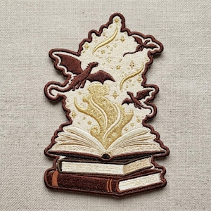 May include: An embroidered patch featuring two stacked books with an open book on top. Golden magical energy rises from the pages, surrounded by two small dark brown dragons, stars, and crescent moon shapes against a cream-colored background.
