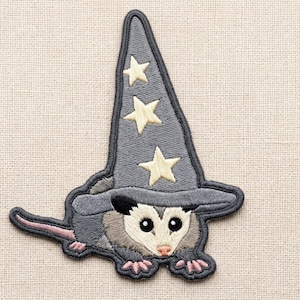 May include: An embroidered patch featuring a gray opossum wearing a tall, pointed gray wizard hat decorated with three cream-colored stars. The opossum has a white face, black eyes, a pink nose, and small pink feet, with a long pink tail extending to the left.