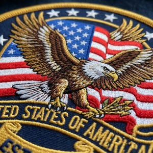 May include: A circular embroidered patch featuring a bald eagle with brown and white feathers, wings spread wide, clutching arrows. The background displays the American flag with red, white, and blue colors. A gold banner reads UNITED STATES OF AMERICA.