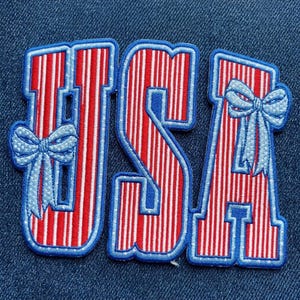 May include: An embroidered patch featuring the letters USA in a red and white vertical striped pattern with a blue border. The letter U is decorated with a light blue bow on the left side, and the letter A features a light blue bow on the right side.