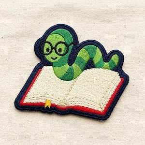 May include: An embroidered patch featuring a green worm wearing black round glasses, resting on an open book with cream-colored pages, a red cover, and a small yellow bookmark, all outlined with a thick dark blue border.