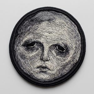 May include: A circular embroidered patch featuring a detailed human face rendered in shades of gray, white, and black thread. The face has deep-set eyes, a prominent nose, and a neutral mouth, all enclosed within a thick, solid black stitched border.