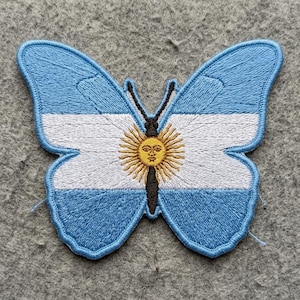 May include: An embroidered butterfly-shaped patch featuring the flag of Argentina. The design displays horizontal light blue and white stripes with a central yellow sun with a human face, all outlined in a thick, dark blue stitched border.
