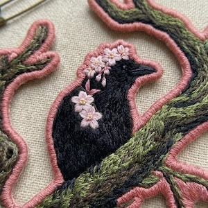 May include: An embroidered patch featuring a black bird perched on a textured green and brown tree branch. The bird is adorned with small, light pink cherry blossom flowers. The entire design is outlined with a thick, raised pink thread border.