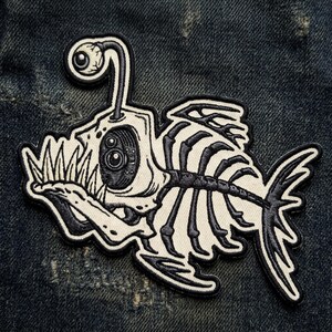 May include: An embroidered patch featuring a skeletal anglerfish design in black and off-white thread. The fish has a prominent lure with an eyeball on top, sharp teeth, a large circular eye, and a visible ribcage structure with a fin and tail.