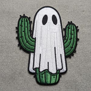May include: An embroidered patch featuring a green cactus wearing a white ghost costume. The ghost has two black oval eyes and is draped over the cactus, which has two arms extending outward. The entire design is outlined in a thick black border.