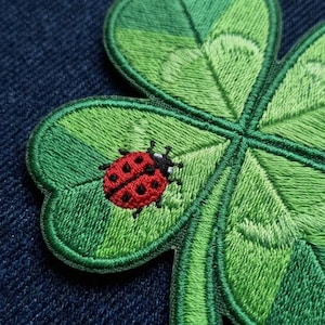 May include: A four-leaf clover patch featuring embroidered green thread in varying shades to create depth and texture. A small, bright red ladybug with black spots is stitched onto one of the clover leaves, providing a distinct color contrast.