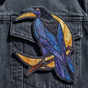 May include: An embroidered patch featuring a stylized bird with blue and purple plumage perched on a branch against a large, golden-yellow crescent moon. The design uses dark thread outlines to create a stained-glass effect on a black denim fabric.