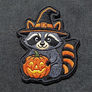 May include: An embroidered patch featuring a gray raccoon wearing a black witch hat with an orange band and buckle. The raccoon is holding a bright orange jack-o'-lantern pumpkin with a carved face. The patch has a black border and striped tail.