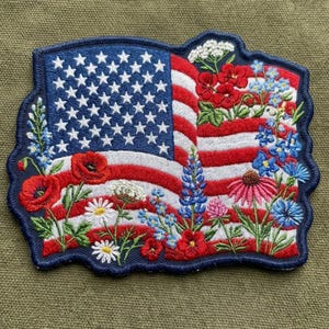 May include: An embroidered patch featuring the American flag with a wavy design, overlaid with a variety of colorful wildflowers including red poppies, blue lupines, white daisies, and pink coneflowers, all bordered by a dark blue stitched edge.