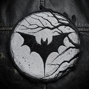 May include: A circular embroidered patch featuring a black bat silhouette centered against a light gray moon. The design includes dark, bare tree branches extending across the moon's surface, all enclosed within a thick black stitched border.