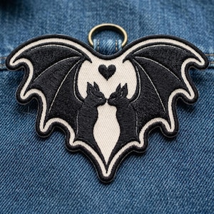 May include: A heart-shaped embroidered patch featuring two black bats facing each other with their wings spread wide against a cream-colored background. A small black heart is positioned between the bats' heads, and a gold metal ring is attached at the top.