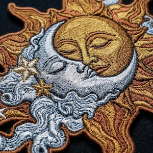 May include: An embroidered patch featuring a golden sun with a human face and closed eyes overlapping a silver crescent moon with a human face. The design includes swirling silver clouds and small golden stars against a dark background.