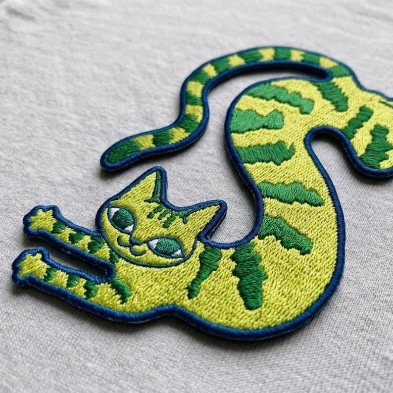 May include: An embroidered patch shaped like a long, flexible cat with yellow and green striped fur. The cat has a dark blue stitched border, large green eyes, and is depicted in a curved, stretching pose with its front paws extended forward.